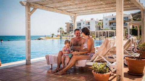 Two parents and a toddler resting in the shade of a beach pergola at midday